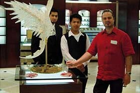 This is an image featuring three individuals standing in a showroom. The focal point of the image appears to be a large, intricate glass sculpture that resembles a bird with outstretched wings, positioned on a display stand. The man wearing a red shirt is gesturing towards the sculpture, possibly explaining its design or craftsmanship. Behind him and slightly to the side is another man in a dark-colored suit looking at the sculpture with interest, while a woman in a light-colored top stands behind both of them, also observing the artwork. The showroom itself has elegant lighting fixtures overhead and what looks like display cases that could be used for showcasing glassware or other delicate items. The setting suggests this might be an event or a store specializing in high-quality glass art, possibly in Venice, Italy, which is famous for its Murano glass craftsmanship. Keywords: MURANO GLASS, VASE, LAMP, MURANO GLASS, DESIGN, MADE IN ITALY, VENICE GLASS