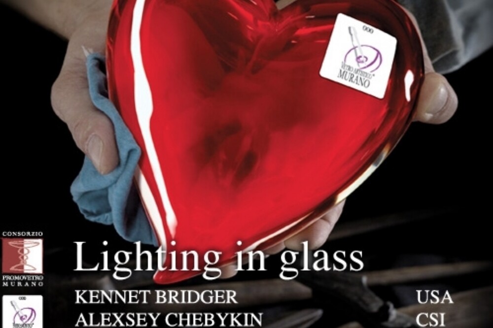 The image features a heart-shaped object that appears to be crafted from molten glass. It's being held by someone who seems to be either shaping the glass on a metal rod or pulling it from a furnace, which is common in traditional glassmaking techniques such as those used in Murano, Italy. The background suggests an industrial setting, possibly a workshop dedicated to creating blown glass objects. The heart itself has a deep red color and looks smooth, indicating that it's either freshly made or has been finished with great care. There are also some white text elements overlaid on the image: 