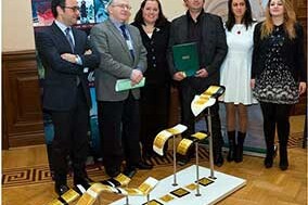 This image depicts a group of people standing around a display that features what appears to be a collection of Murano glass products. The individuals are dressed in formal business attire, suggesting they may be attending an event or meeting related to the glass industry. The man on the right side is holding a plaque with some text and a ribbon, which typically signifies an award or recognition for achievement. The background includes a banner with the name 