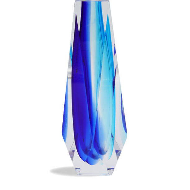 The image shows a tall, slender vase with an elegant design. It features a blend of blue hues that create a striking pattern, reminiscent of flowing water or abstract designs. The vase has a clear body with the blue tinted design more concentrated towards the top and fading towards the bottom, which gives it a dynamic look as if the colors are cascading down its sides. This piece is made from glass, likely originating from Venice given the traditional technique that is associated with Venetian glassware. The craftsmanship appears to be high-quality, suggesting that it could be a work of art or an expensive decorative item. The vase stands alone against a neutral background, which allows its colors and design to stand out prominently. The term 