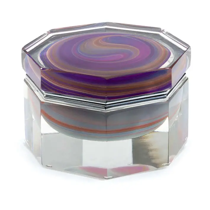 The image shows a piece of glass art that appears to be an ottoman or stool. The top is made from multiple layers of glass, with swirling patterns in shades of purple and magenta, giving it a marbled effect. These layers are encased within a clear glass frame, which provides structural support and enhances the reflective qualities of the piece. The glass design is indicative of Murano glass, which is a type of glassware made on the Venetian island of Murano in Italy. It's known for its fine craftsmanship, including techniques like this layered, swirling effect, often referred to as 