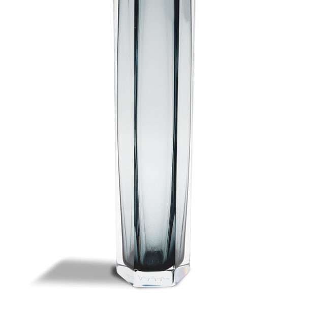 The image shows a tall, slender glass vase with vertical ribbing on its exterior. The glass appears to be of the Murano style, which is known for its intricate designs and craftsmanship. The design suggests that it could be an item created by the Alessandro Mandruzzato studio, as indicated in your description. The vase has a reflective quality, indicating it's made from a type of glass that refracts light to some extent. It is placed on a surface with a neutral background, which puts the focus on the vase itself. The craftsmanship and the aesthetic suggest that this is a piece designed for home decor or as an artisanal creation, likely made in Italy given the style's historical association with Venetian glassblowing traditions.