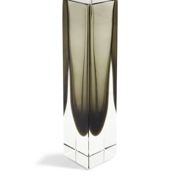 The image shows a tall, slender Murano glass vase with a vertical ribbed design and a slightly tapered shape that narrows towards the bottom. The color of the vase appears to be a dark green or teal hue with subtle variations in tone throughout its length. It's placed on a simple stand against a neutral background, which highlights the piece's form and texture. Murano glass is renowned for its artistry and craftsmanship, often characterized by intricate designs and vibrant colors. The vase's sleek profile suggests contemporary design, while the ribbing adds an organic, almost cellular pattern to its surface.