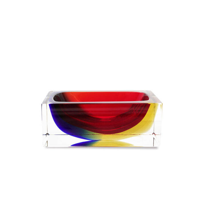 The image features a striking piece of Murano glass art. The glass appears to be crafted into an abstract bowl-like form, with a vibrant design that includes shades of red, yellow, and blue. The colors are layered in a way that gives the impression of depth and movement, suggesting a dynamic interplay between light and shadow. The bowl is placed on a clear stand, which contrasts with the rich colors of the glass and allows the piece to be seen from below as well as above. This placement also enhances its prominence and makes it look like a centerpiece. The background is neutral, ensuring that all attention remains on the glass artwork. The style of the image is minimalist, focusing solely on the glass piece without any distractions. This approach highlights the craftsmanship and artistic value of the Murano glass, emphasizing its qualities as an object of design and beauty.