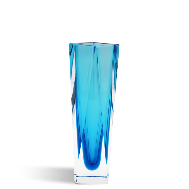 The image shows a tall, elegant glass vase with an intricate design. The vase is predominantly blue in color with varying hues that create a sense of depth and movement. Its form includes swirling patterns reminiscent of ocean waves or the flow of water, which gives it a dynamic and fluid appearance. This piece appears to be handcrafted, as suggested by the uneven edges and organic shapes within the design. The vase is standing on a flat surface, and there's a subtle reflection beneath it, indicating that the surface might be reflective or polished. The background is neutral-toned with a gradient from light at the top to slightly darker at the bottom, which helps to highlight the blue of the glass vase. There are no texts visible in the image. The style of the vase suggests it could be Murano glassware, known for its artistic designs and traditional techniques associated with Venice, Italy.