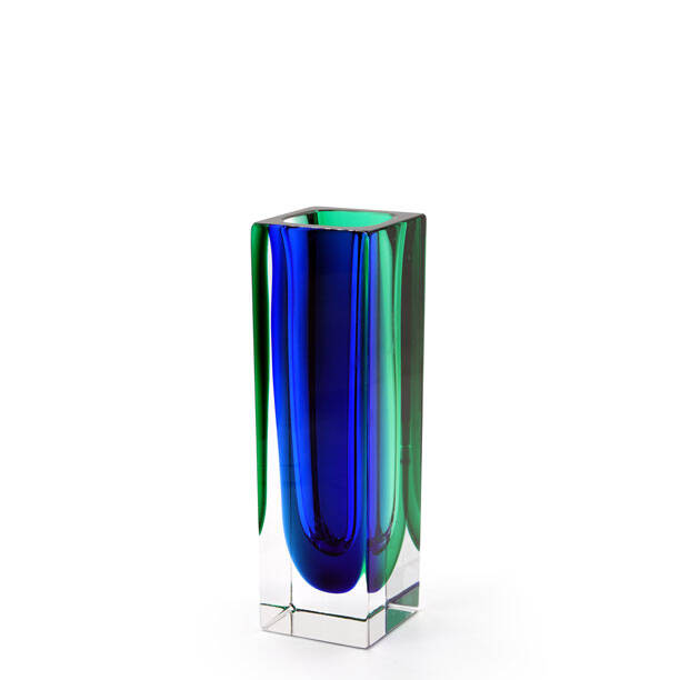 The image shows a tall, slender glass vase with a colorful design that transitions from green at the top to blue and then to a darker green or black towards the bottom. The colors appear smooth and are likely achieved through a layering technique in which different colored layers of molten glass are blown onto each other. The vase is placed on a clear stand, possibly for display purposes, against a neutral background that contrasts with the vibrant colors of the glass piece. The craftsmanship suggests it could be an example of Murano glass, known for its intricate and colorful designs originating from Venice, Italy.