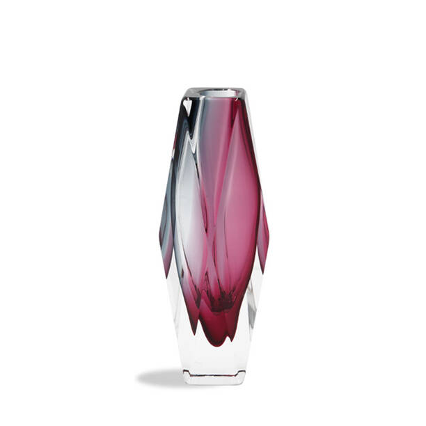 This image showcases a beautifully crafted vase made from Murano glass. The vase has an elongated form with a slender neck that tapers slightly towards the bottom, giving it a graceful and elegant silhouette. The color of the vase is a rich, deep purple, which adds a touch of sophistication to its appearance. The craftsmanship of the Murano glass is evident in the subtle variations in the color depth throughout the vase, suggesting an artisanal process where individual elements are handcrafted and blended together. These variations create a mesmerizing effect that captures light in different ways, adding a dynamic aspect to the piece. The background of the image has a gradient that goes from a pale pink at the top to white towards the bottom, which helps to highlight the vase as the focal point. There's also a subtle shadow beneath the vase, indicating that it is placed on a surface, possibly a table or a stand. The text 