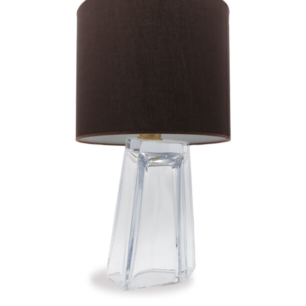 The image shows a modern table lamp with a distinctive design. The base of the lamp is transparent, resembling glass in its structure but potentially made from a different material like acrylic or plastic, giving it an elegant and contemporary look. It has an unusual shape that could be compared to the bottom part of a vase or a glass carafe, with what appears to be ridges or facets along its sides, which adds depth and texture. The lampshade is cylindrical and seems to be made from a fabric-like material, possibly felt or velvet, in a dark brown color that contrasts nicely with the transparency of the base. The shade is attached directly to the base without any visible support, giving it a seamless appearance. In terms of branding or provenance, there's no text present on the image itself, but if this lamp were associated with 