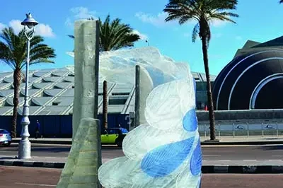 The image depicts a scene that appears to be in front of a Murano glass factory. The focal point is an art installation or sculpture made from large, colorful glass pieces arranged in a manner reminiscent of a flower or abstract plant form. It's situated on the street near what seems to be a palm tree-lined road. In the background, there are buildings that could possibly be part of the factory complex, and beyond them, you can see a modern structure with a distinctive architectural design that stands out from the more traditional surroundings. The sky is clear, suggesting it might be a warm or summer day.