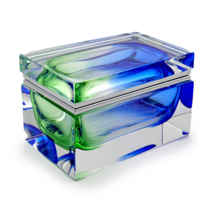 The image shows a piece of Murano glass, which is renowned for its intricate designs and vibrant colors. The object appears to be a rectangular container with rounded corners. It has a layered design that incorporates multiple colors; the top layer seems to have a mix of blue tones while the lower layers transition through green shades, giving it an iridescent effect. The Murano glass style is characterized by its handcrafted quality and the use of traditional Venetian techniques like lampworking or blown glass methods. The craftsmanship involves precise control over the heating and shaping processes to achieve such layered and smooth transitions between colors. This particular piece could be used as a decorative object, perhaps for holding jewelry, keys, or other small items due to its box-like structure. It's an example of Venetian glass artistry that is highly sought after by collectors and enthusiasts of fine glassware.