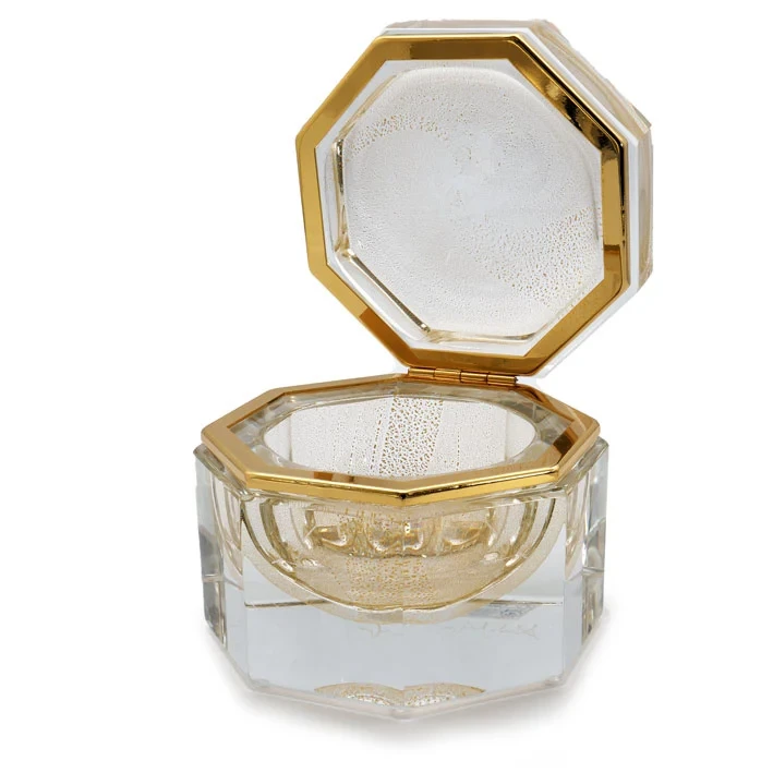 The image shows a Murano glass product, which is known for its intricate designs and craftsmanship. This particular piece appears to be a small perfume bottle with an attached lid that has a honeycomb-like pattern on the top. The body of the bottle is clear, allowing one to see inside, while the base features gold accents, adding a touch of elegance and sophistication to the overall design. Murano glass is made in Italy, specifically on the island of Murano near Venice, and it's renowned for its vibrant colors and delicate artistry that often includes patterns, designs, and decorative elements crafted by skilled glassblowers.