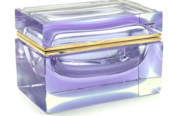 The image shows a beautifully crafted piece of Murano glass, specifically a box with an intricate design. The box has a deep purple hue that appears to be swirled or marbled throughout its structure, creating a mesmerizing effect. There's a gold-colored band around the center that contrasts with the vibrant color and adds a touch of elegance to the piece. Murano glass is renowned for its artistry and craftsmanship, and this box is no exception. The design suggests it could be used as a decorative object or perhaps as a jewelry box given its size and the attention to detail in its construction. The image showcases the skill involved in creating such glassware, emphasizing the beauty of Italian craftsmanship.