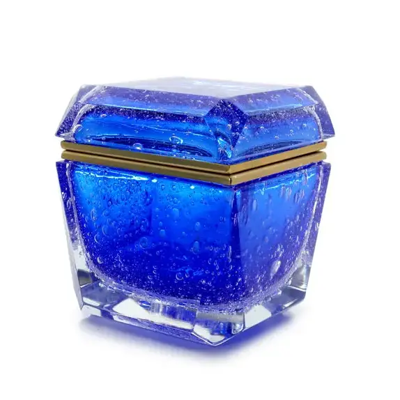 The image shows a small, decorative glass container with a blue tinted interior. The exterior of the lid is smooth and also has a blue color, but it seems to be a solid shade rather than translucent like the inside. The base of the container appears to have a clear section that allows for visibility into the compartment, which might suggest it's designed to hold small items such as rings or other jewelry. The design and quality of craftsmanship indicate that this item could be made by 