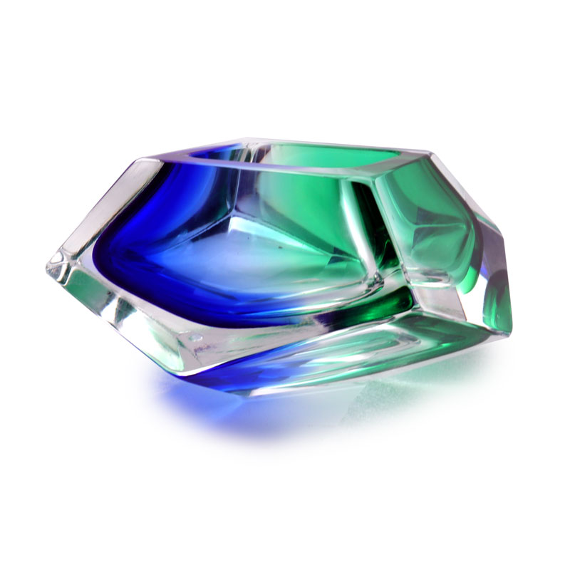 The image features a striking piece of Murano glass art. The glass appears to be crafted into an abstract bowl-like form, with a vibrant Cobalt and green color. The colors are layered in a way that gives the impression of depth and movement, suggesting a dynamic interplay between light and shadow. The bowl is placed on a clear stand, which contrasts with the rich colors of the glass and allows the piece to be seen from below as well as above. This placement also enhances its prominence and makes it look like a centerpiece. The background is neutral, ensuring that all attention remains on the glass artwork. The style of the image is minimalist, focusing solely on the glass piece without any distractions. This approach highlights the craftsmanship and artistic value of the Murano glass, emphasizing its qualities as an object of design and beauty.